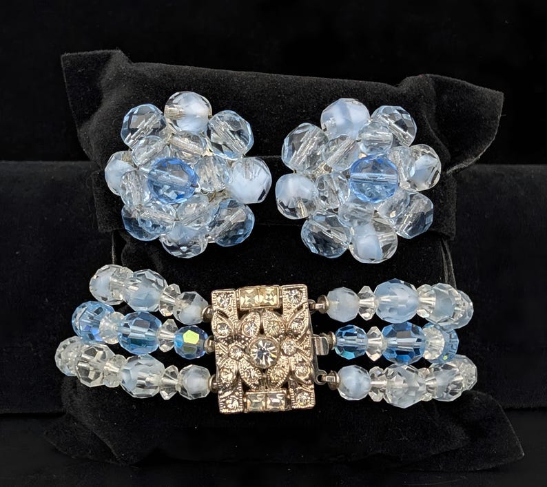 May include: A vintage jewelry set featuring a bracelet and matching earrings. The bracelet has three strands of clear and light blue beads with a decorative silver clasp. The earrings are clusters of similar beads. The set is displayed on a black velvet surface.
