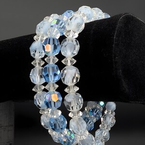 May include: A three-strand bracelet featuring faceted beads in shades of light blue and clear. The beads are strung together with clear spacers, creating a delicate and sparkling accessory. The bracelet is displayed against a black background.