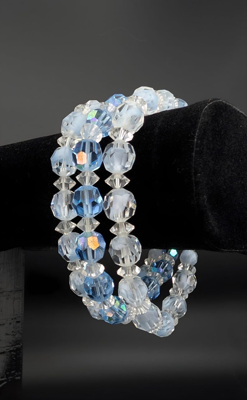 May include: A three-strand bracelet featuring faceted beads in shades of light blue and clear. The beads are strung together with clear spacers, creating a delicate and sparkling accessory. The bracelet is displayed against a black background.