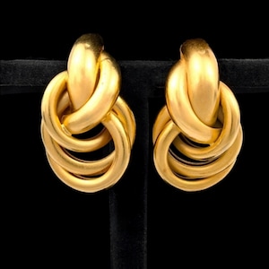 May include: A pair of gold-toned earrings with a knotted design. The earrings have a polished finish and a three-dimensional, intertwined loop structure. The background is black, highlighting the earrings' metallic sheen.