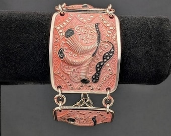 May include: A rectangular bracelet featuring a salmon-pink enameled surface with intricate embossed scrollwork and black accents. The piece includes a smaller matching rectangular link connected by metal rings, showcasing a detailed textured design.