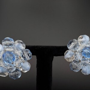 May include: A pair of vintage clip-on earrings. Each earring features a cluster of clear and light blue faceted beads, with a larger blue bead at the center. The earrings are displayed on a black stand against a dark background.