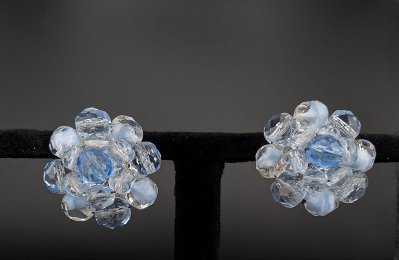 May include: A pair of vintage clip-on earrings. Each earring features a cluster of clear and light blue faceted beads, with a larger blue bead at the center. The earrings are displayed on a black stand against a dark background.
