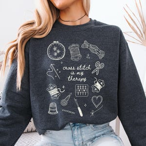 May include: Dark gray sweatshirt with white line art of cross-stitch tools and the text "cross stitch is my therapy." The design includes a needle, thread spools, scissors, and a heart. The sweatshirt is a comfortable, casual garment.
