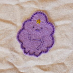 May include: A purple felt patch in the shape of a cloud with a smiling face and a gold star. The cloud has a dark purple stitched outline and embroidered details. The patch is on a beige fabric background.