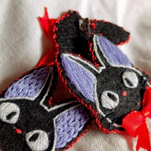 Puede incluir: Par de broches de fieltro con forma de gato negro con orejas bordadas en lavanda y detalles blancos. Los broches tienen bordes cosidos en rojo y pequeños lazos rojos. El fondo es de tela blanca.