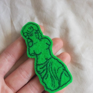 Può includere: Applicazione in feltro verde a forma di statua classica. La figura è delineata con filo nero, mostrando i dettagli della forma. L'applicazione è tenuta in una mano.