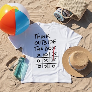 Może przedstawiać: Biała koszulka z napisem "THINK OUTSIDE THE BOX" i grą w kółko i krzyżyk. Piłka plażowa, butelka na wodę, słomkowy kapelusz i tkana torba znajdują się na piaszczystej powierzchni.
