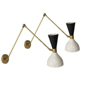 May include: Two mid-century modern wall sconces. Each sconce features a white, bell-shaped shade with black accents and a brass-toned adjustable arm. The shades have small, evenly spaced holes. The sconces are mounted on round brass-toned wall plates.