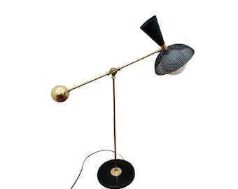 May include: A mid-century modern floor lamp with a black and white perforated shade and a black conical shade. The lamp features a brass-colored arm and base, and a gold sphere on the end of the arm. The lamp is plugged into a wall outlet.