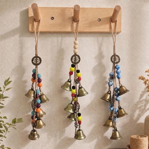 Peut inclure: Trois décorations murales suspendues à un support en bois clair à trois patères. Chaque ornement comporte une breloque pentagramme en bronze, des clochettes en métal et des perles colorées orange, bleu, jaune, rouge et noir, avec des feuilles.