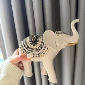 May include: A white elephant figurine with a decorative saddle featuring black and gold patterns. The elephant has a raised trunk and large ears. The figurine is held against a gray curtain backdrop.