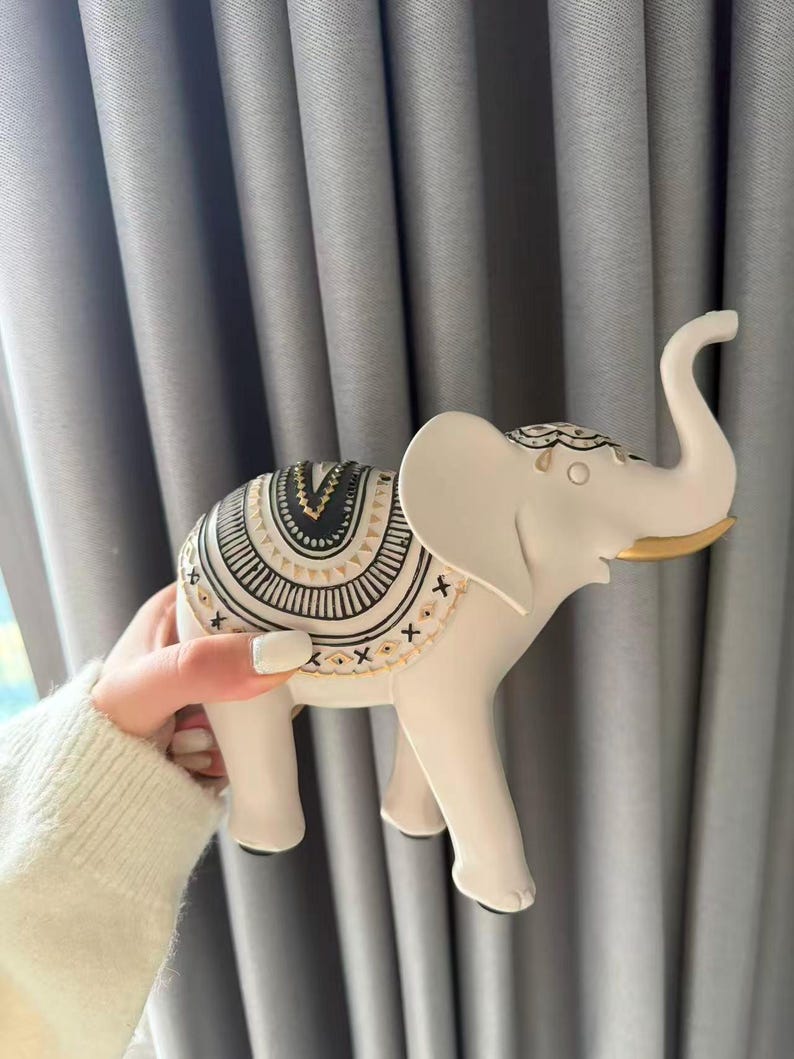 May include: A white elephant figurine with a decorative saddle featuring black and gold patterns. The elephant has a raised trunk and large ears. The figurine is held against a gray curtain backdrop.