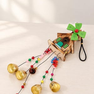 Peut inclure: Un ornement décoratif en bois en forme de moulin à vent avec une structure de maison marron et une hélice verte, agrémenté de cordons suspendus de perles colorées, de breloques papillons et de quatre clochettes en métal doré brillant.