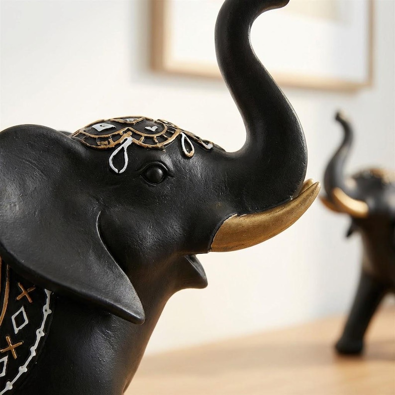 May include: A black elephant figurine with gold accents on the tusks and decorative headpiece. The elephant's trunk is raised, and the figurine has a detailed, textured finish. A second elephant figurine is slightly out of focus in the background.