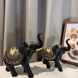 May include: Two black elephant figurines with gold and white decorative patterns. The elephants have raised trunks and are positioned on a white surface. The larger elephant is in the foreground, with a smaller one behind it.