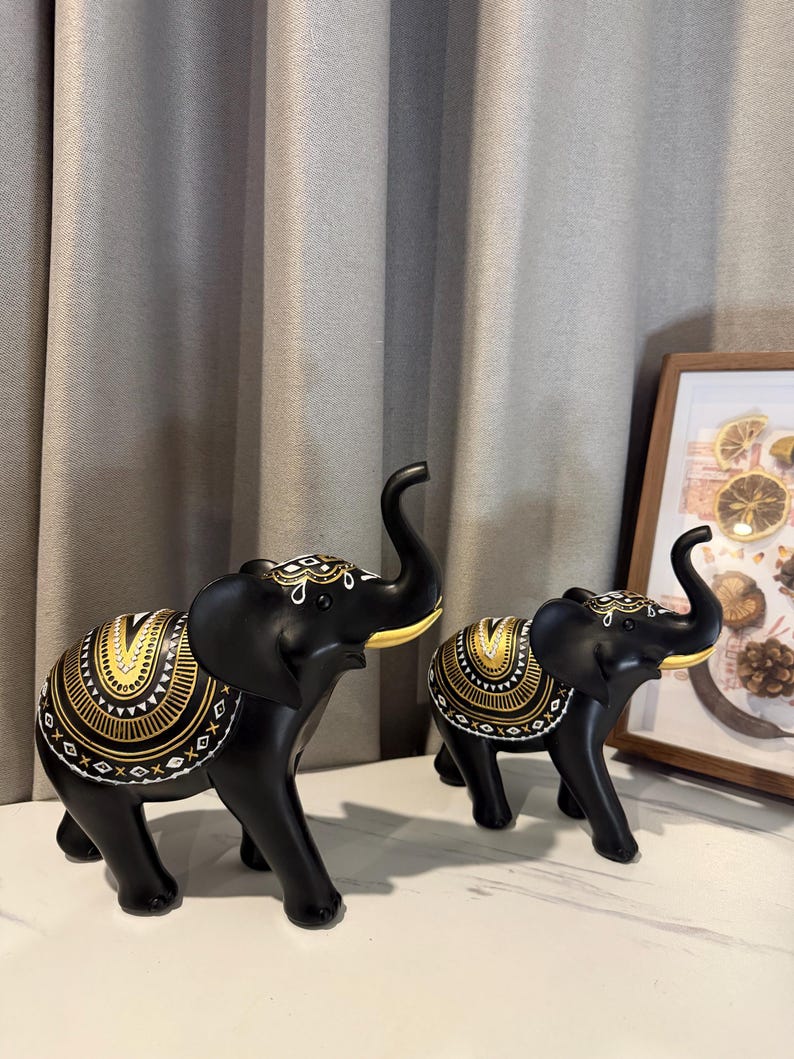 May include: Two black elephant figurines with gold and white decorative patterns. The elephants have raised trunks and are positioned on a white surface. The larger elephant is in the foreground, with a smaller one behind it.