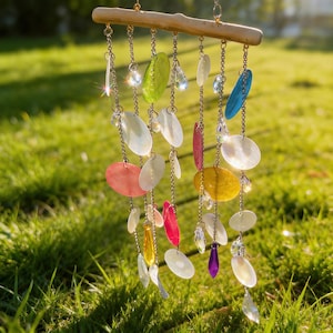 Puede incluir: Un carillón de viento decorativo con piezas coloridas en forma de concha iridiscente y detalles de cristal. El carillón cuelga de una rama de madera natural y está suspendido por delicadas cadenas. Los colores incluyen rosa, amarillo, verde y azul.