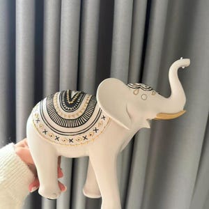 May include: A white elephant figurine with intricate black and gold patterns on its back. The elephant has its trunk raised and gold accents on its ears. The figurine is a decorative item.