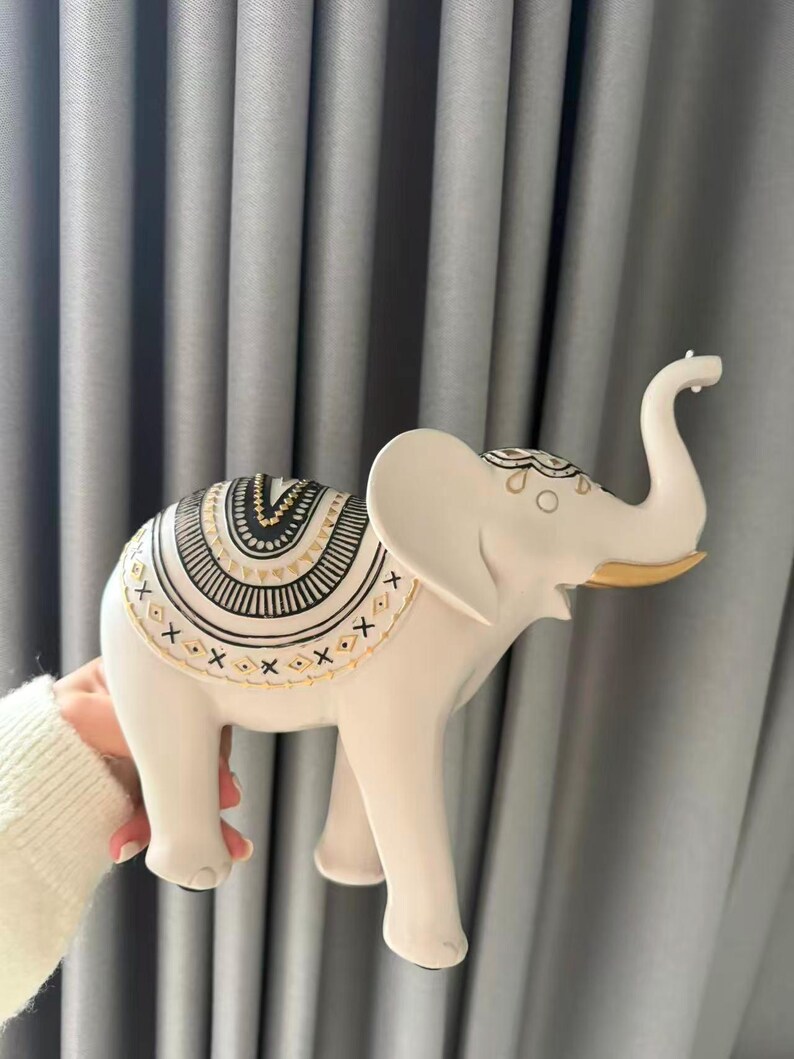 May include: A white elephant figurine with intricate black and gold patterns on its back. The elephant has its trunk raised and gold accents on its ears. The figurine is a decorative item.