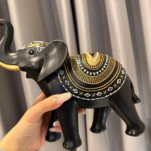 May include: A black elephant figurine with gold and white decorative accents. The elephant has a raised trunk and detailed patterns on its back and head. The figurine is held by a hand, showcasing its size and design.