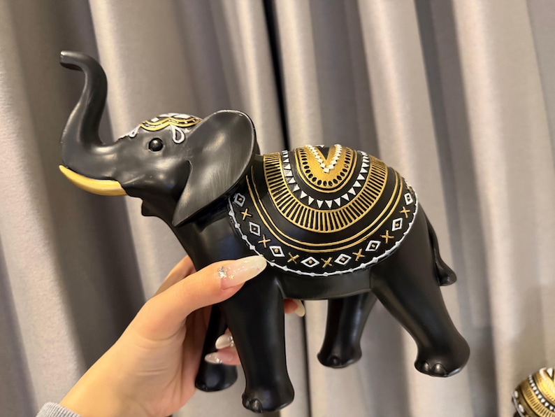 May include: A black elephant figurine with gold and white decorative accents. The elephant has a raised trunk and detailed patterns on its back and head. The figurine is held by a hand, showcasing its size and design.