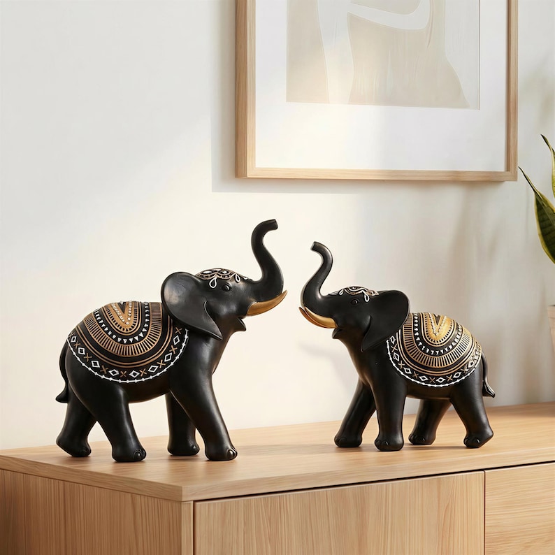 May include: Two black elephant figurines with gold and white decorative patterns on their backs. The elephants have raised trunks and are positioned facing each other. The figurines are displayed on a wooden surface.