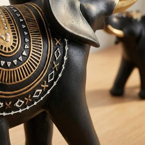May include: Close-up of a black elephant figurine with gold accents. The elephant has a decorative saddle with intricate gold and white patterns. The figurine is on a wooden surface.