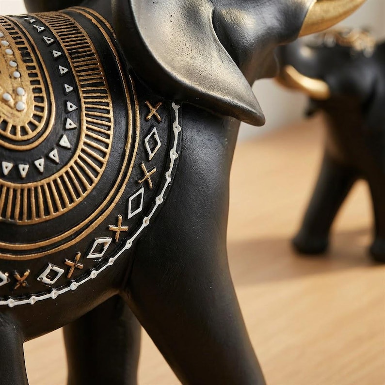 May include: Close-up of a black elephant figurine with gold accents. The elephant has a decorative saddle with intricate gold and white patterns. The figurine is on a wooden surface.