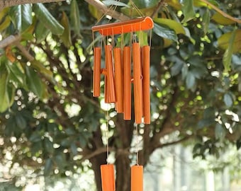 May include: A hanging wind chime with orange-brown bamboo tubes and a matching top piece. The chimes are suspended from a tree branch, with a blurred green background of leaves and branches.
