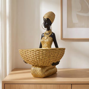 May include: A decorative figurine of a woman with black skin, wearing gold jewellery and a woven headdress and dress. The figurine holds a large, woven basket. The statue is on a wooden surface.