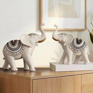May include: Two ivory-colored elephant figurines with raised trunks and decorative saddle blankets. The blankets feature black, gold, and white geometric patterns. The elephants are displayed on a wooden surface.