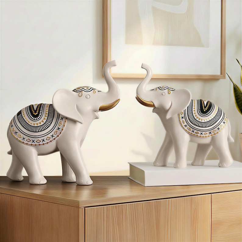 May include: Two ivory-colored elephant figurines with raised trunks and decorative saddle blankets. The blankets feature black, gold, and white geometric patterns. The elephants are displayed on a wooden surface.