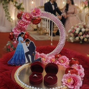 May include: A circular ring display tray featuring a pink ribbon-wrapped hoop adorned with pink and red artificial flowers, pearl accents, and a cutout of a couple. Two heart-shaped velvet ring boxes hold rings on a reflective silver base.