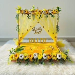 Op de afbeelding: Een decoratieve gele Haldi-tray met bloemstukken en de tekst "Shailly Jay" en "HALDI". De tray is versierd met gele stof, parels en een verscheidenheid aan gele en witte bloemen, wat een feestelijke uitstraling creëert.