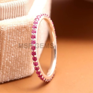 May include: A silver ring with a band of small, vibrant red gemstones. The ring is set against a soft, beige velvet jewelry box. The ring is a delicate piece of jewelry, perfect for everyday wear or special occasions.