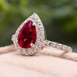 Puede incluir: Un anillo con una piedra preciosa roja rubí en forma de pera, rodeada por un halo de pequeñas piedras claras. La banda también está adornada con pequeñas piedras claras, engastadas en un metal plateado. El anillo se muestra sobre una superficie de tono neutro.