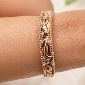 May include: A rose gold ring featuring an intricate engraved floral pattern along the band, bordered by delicate beaded edges. The metal has a warm, metallic sheen, and the design consists of repeating curved leaf-like motifs that wrap around the entire circumference of the band.