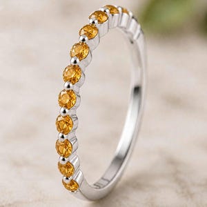 Puede incluir: Un anillo de banda en tono plateado que presenta una hilera de pequeñas piedras preciosas de citrino amarillo, redondas y facetadas, engastadas en un estilo de garras compartidas. La banda metálica tiene un acabado pulido y las piedras.