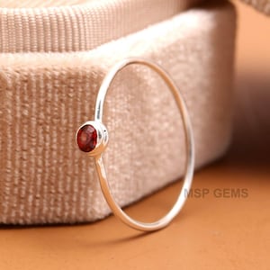 Puede incluir: Un delicado anillo de plata con una pequeña gema redonda de color rojo intenso. El anillo tiene una banda fina y un diseño simple y elegante. La gema está engastada en un bisel.