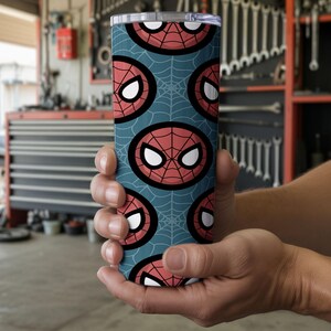Peut inclure: Un gobelet cylindrique avec un motif r&eacute;p&eacute;t&eacute; du masque de Spider-Man sur un fond bleu sarcelle avec des toiles d'araign&eacute;es. Le gobelet est tenu dans les mains d'une personne. Le gobelet a un couvercle argent&eacute;.