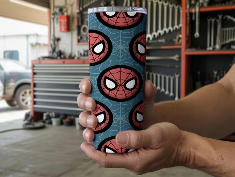 Peut inclure: Un gobelet cylindrique avec un motif r&eacute;p&eacute;t&eacute; du masque de Spider-Man sur un fond bleu sarcelle avec des toiles d'araign&eacute;es. Le gobelet est tenu dans les mains d'une personne. Le gobelet a un couvercle argent&eacute;.