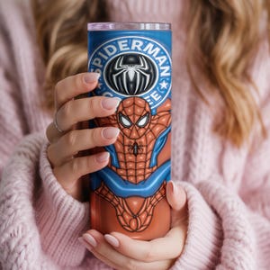 May include: A tall, cylindrical tumbler featuring a Spider-Man design. The tumbler is primarily blue and orange, with the Spider-Man logo at the top. The design includes the Spider-Man character in a dynamic pose. The tumbler is held by a person wearing a pink sweater.