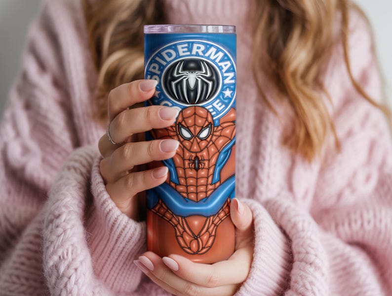 May include: A tall, cylindrical tumbler featuring a Spider-Man design. The tumbler is primarily blue and orange, with the Spider-Man logo at the top. The design includes the Spider-Man character in a dynamic pose. The tumbler is held by a person wearing a pink sweater.