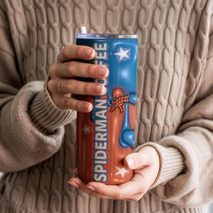 May include: A stainless steel tumbler featuring a design with the words "SPIDERMAN COFFEE" in a vertical layout. The tumbler has a blue and red color scheme with star accents and a graphic of Spider-Man.