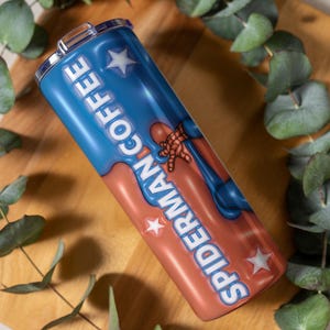 May include: A stainless steel tumbler with a blue and red design featuring the words "COFFEE" and "SPIDERMAN" in white. The tumbler has a silver lid and is decorated with white stars and a Spiderman graphic.