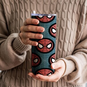Peut inclure: Un gobelet cylindrique et haut avec un motif r&eacute;p&eacute;t&eacute; du masque de Spider-Man en rouge et blanc sur un fond bleu sarcelle avec un motif de toile d'araign&eacute;e. Le gobelet est tenu par une personne portant un pull beige.
