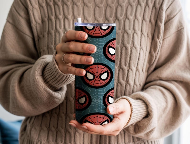 Peut inclure: Un gobelet cylindrique et haut avec un motif r&eacute;p&eacute;t&eacute; du masque de Spider-Man en rouge et blanc sur un fond bleu sarcelle avec un motif de toile d'araign&eacute;e. Le gobelet est tenu par une personne portant un pull beige.