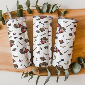 May include: Three white cylindrical tumblers featuring a repeating pattern of cartoon characters riding broomsticks, brown books, golden winged balls, black keys, and yellow lightning bolts. Each tumbler has a metallic rim and a smooth finish.