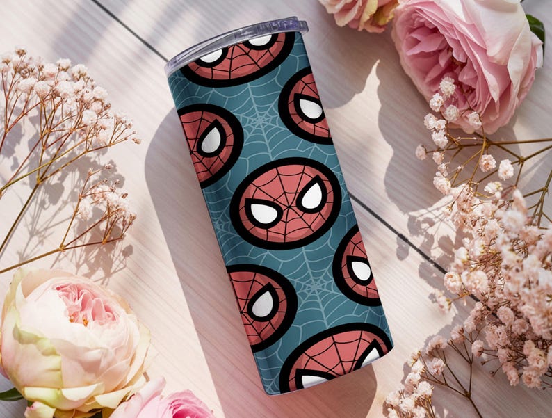 Peut inclure: Un gobelet en acier inoxydable avec un motif r&eacute;p&eacute;t&eacute; du masque de Spider-Man sur un fond bleu sarcelle avec des toiles d'araign&eacute;es blanches. Le gobelet a un couvercle transparent. Entour&eacute; de roses roses et de fleurs blanches.